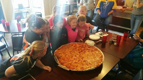 A group of excited children gather around a giant pizza on a table, smiling and eager to dig in.