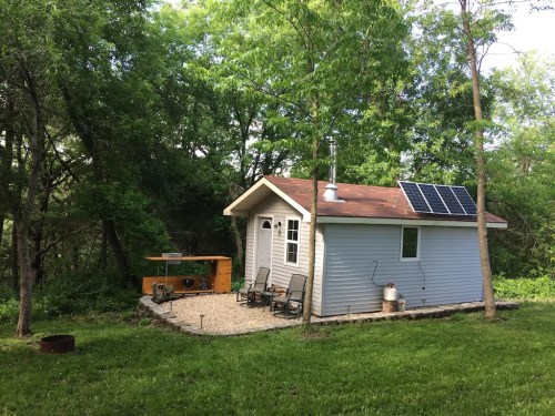 A small, light blue cabin surrounded by trees, featuring a solar panel and a gravel patio with outdoor seating.