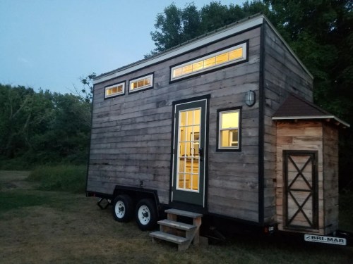 A rustic tiny house on wheels with a wooden exterior, featuring large windows and a small porch, set in a grassy area.