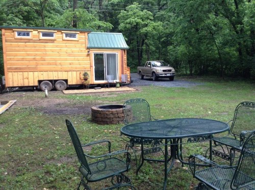 A tiny wooden house on wheels parked in a green, wooded area, with a gravel driveway and a fire pit nearby.