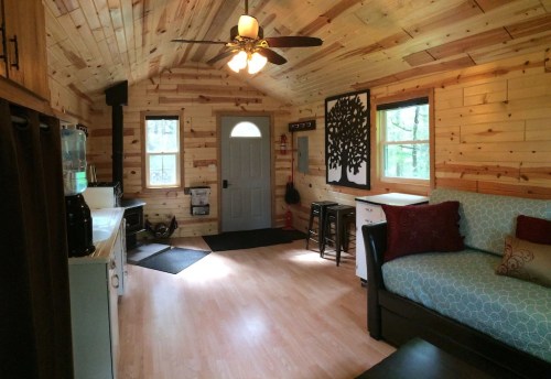 Cozy interior of a cabin with wooden walls, a door, a couch, and a small kitchen area. Natural light filters in.
