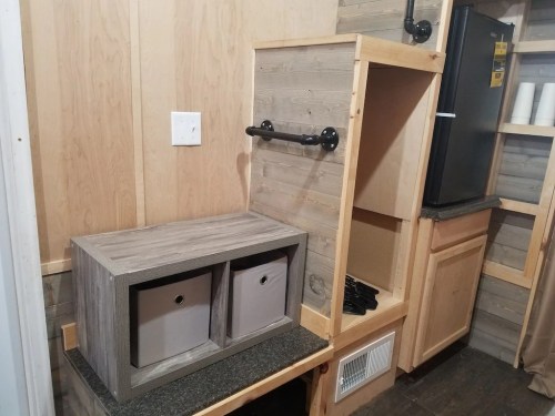 A compact kitchen area featuring a wooden cabinet with two storage bins, a refrigerator, and a shelf with pipes.