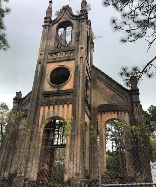 A weathered, abandoned church tower with a broken window, surrounded by trees and a chain-link fence.