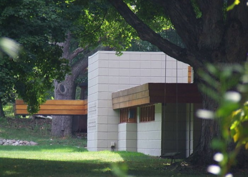 A modern building with a flat roof and wooden accents, surrounded by lush greenery and large trees.
