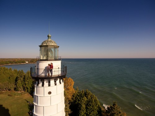 A couple stands on the balcony of a lighthouse overlooking a calm lake and lush green trees under a clear blue sky.