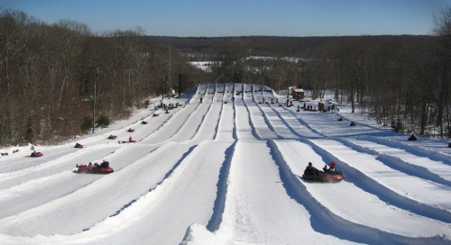 A snowy hill with multiple tubing lanes, people sliding down on inflatable tubes under a clear blue sky.