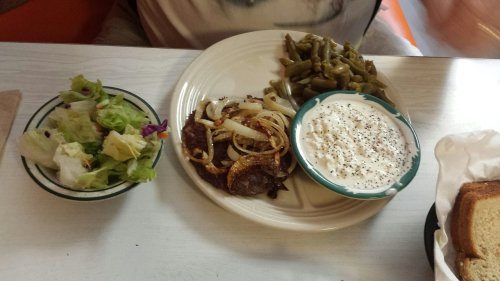 A plate with sautéed onions and meat, green beans, a side salad, and a bowl of creamy dressing on a table.