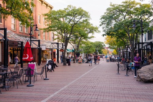 A lively pedestrian street lined with shops and trees, featuring outdoor seating and people enjoying the atmosphere.