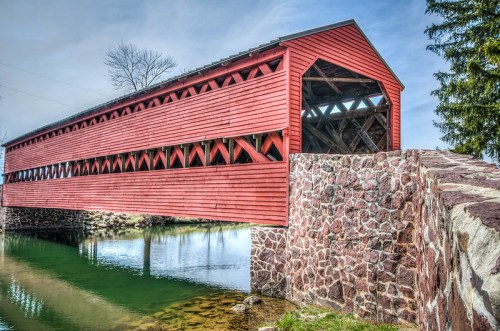 A red covered bridge spans a calm river, with stone supports and trees in the background under a clear sky.