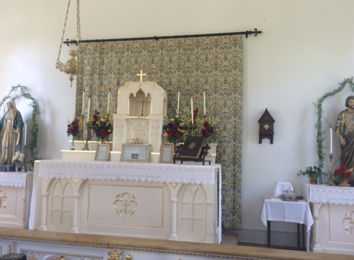 A church altar adorned with flowers, candles, and religious statues, set against a patterned backdrop.