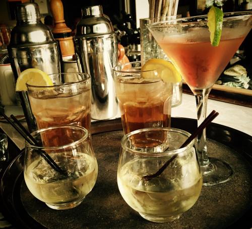 A tray with four drinks: two cocktails with lemon slices and two glasses of clear liquid, surrounded by bar tools.
