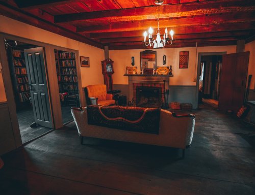 Cozy, dimly lit living room with wooden beams, a fireplace, vintage furniture, and bookshelves in the background.