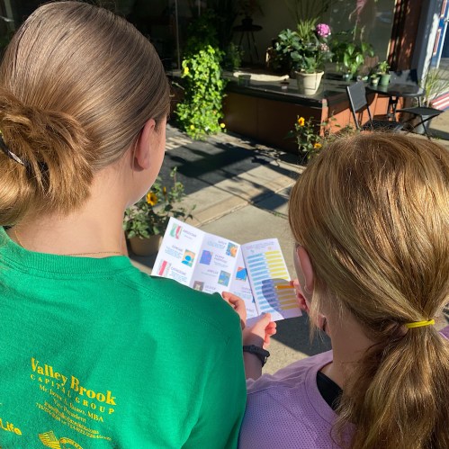 Two children, one in a green shirt and the other in a purple shirt, look at a brochure outside a shop.