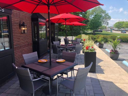 Outdoor dining area with tables under red umbrellas, surrounded by greenery and flowers. Bright and inviting atmosphere.