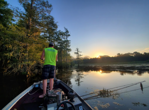 A person fishing from a boat at sunset, surrounded by trees and calm water reflecting the colorful sky.
