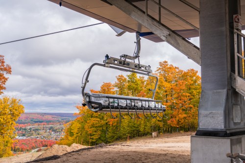 A ski lift chair hangs from a station, surrounded by vibrant autumn trees and a scenic view of the landscape below.