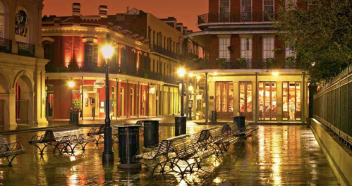 A quiet, illuminated street in a historic district, with benches and wet pavement reflecting warm lights at dusk.