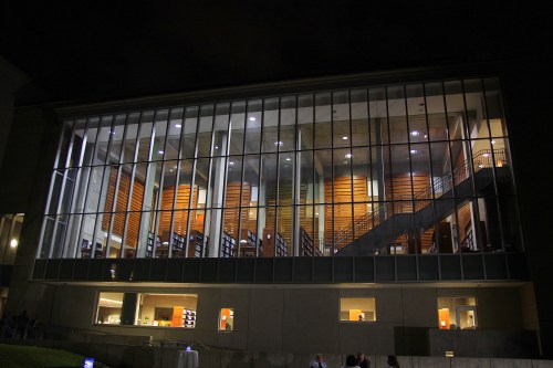 A modern building at night, featuring large glass windows and illuminated interior spaces with stairs and wooden accents.