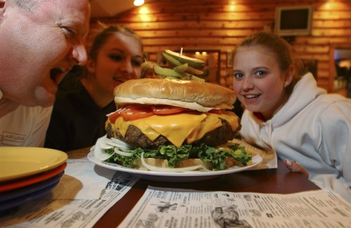 A giant cheeseburger with toppings sits on a table as two girls and a man excitedly look at it.