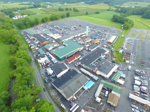 Aerial view of a large market area with multiple buildings, parking lots, and green fields in the background.