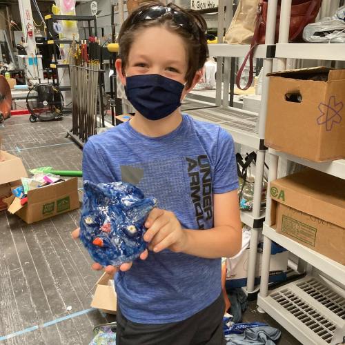 A child wearing a mask holds a blue ceramic owl in a workshop filled with art supplies and boxes.