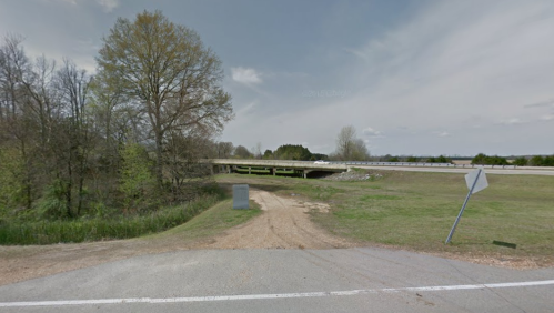 A dirt road leads to an overpass, surrounded by greenery and open fields under a cloudy sky.