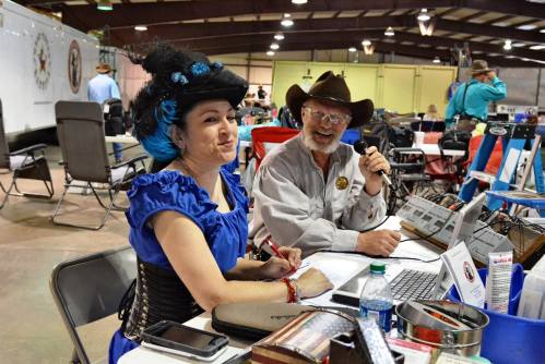 A woman in a blue dress and a man in a cowboy hat sit at a table with microphones and equipment, smiling at the camera.