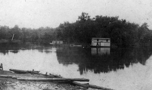 Black and white image of a calm river with a houseboat and trees reflected in the water. Logs are visible in the foreground.