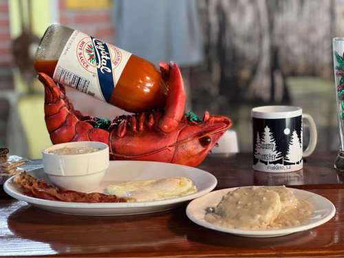 A vibrant lobster-shaped holder with hot sauce, alongside a plate of bacon, eggs, and biscuits with gravy, plus a coffee mug.