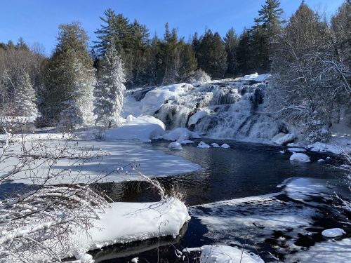 A snowy landscape featuring a waterfall surrounded by trees and a partially frozen river under a clear blue sky.