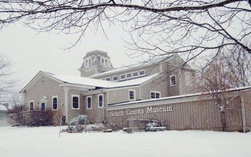Snow-covered South County Museum with a gray exterior and a tower, surrounded by bare trees.