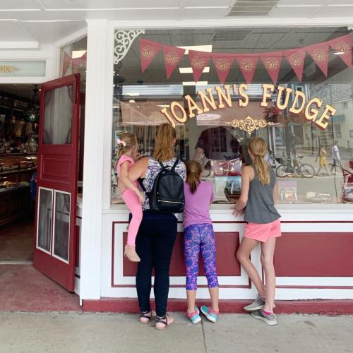 Four children and a woman look at a fudge shop window, with colorful banners and a display of sweets inside.