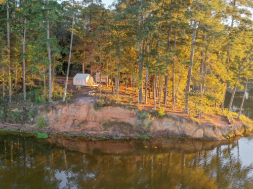 A serene lakeside scene with a tent nestled among tall trees, reflecting on calm water at sunset.