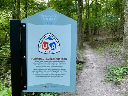 Sign for Bailey Woods Trail, a National Recreation Trail, with a wooded path visible in the background.