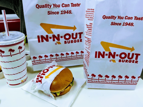 Two In-N-Out Burger bags, a burger on a napkin, and a drink on a table, all featuring the restaurant's logo.