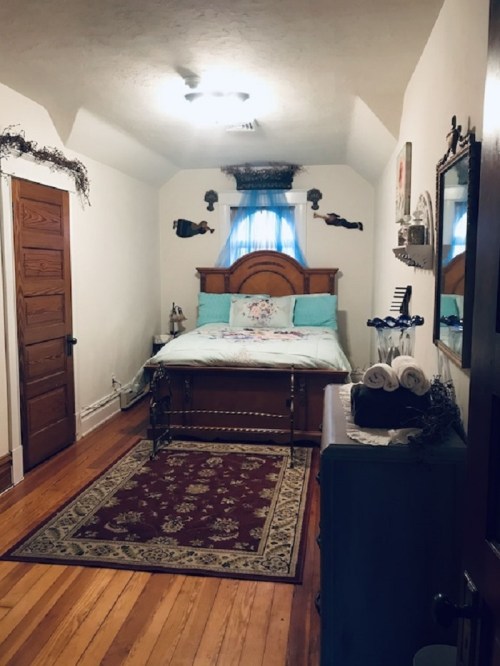 Cozy bedroom with a wooden bed, light blue bedding, a rug, and decorative wall accents. Natural light from a window.