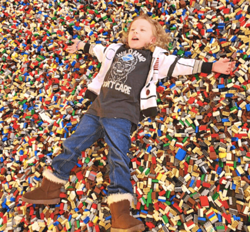 A child lies on a colorful pile of LEGO bricks, arms outstretched, smiling happily.