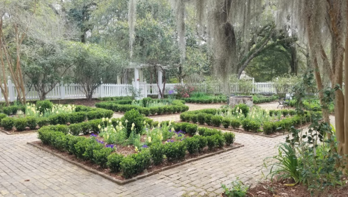 A serene garden with neatly arranged flower beds, surrounded by greenery and a white picket fence.