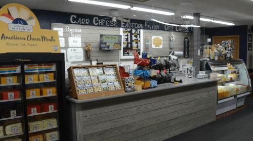 A cheese shop display featuring cheese trays, snacks, and a refrigerated case, with a sign for American Cheese Month.