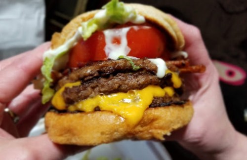 A close-up of a hand holding a juicy burger with two beef patties, cheese, lettuce, tomato, and mayonnaise.