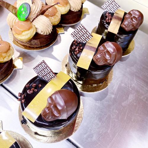 Three chocolate cakes topped with skull decorations and gold accents, alongside cream-filled pastries.