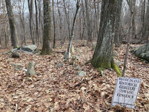 A wooded area with fallen leaves and a sign reading "Rhode Island Historical Cemetery, Town of Lincoln."