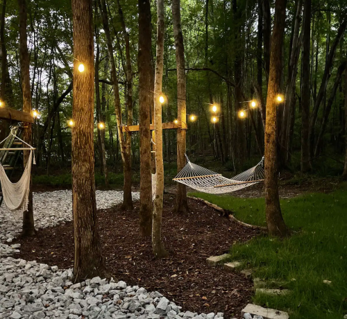 A serene outdoor space with a hammock, string lights, and trees, surrounded by gravel and grass.