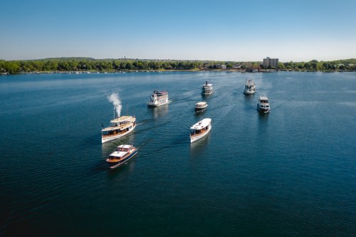 Aerial view of several boats navigating a calm lake on a clear day, surrounded by green trees and buildings.