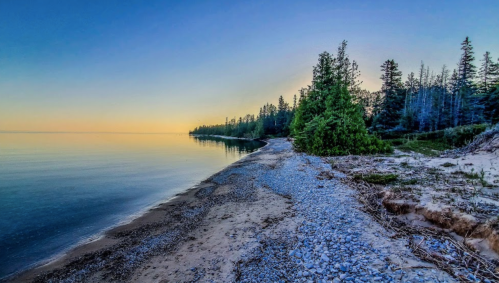 Serene lakeside view at dawn, featuring a pebbly shore and trees against a colorful sky.