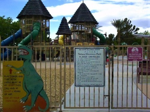 A fenced playground with colorful structures, a dinosaur mural, and a sign detailing park rules.