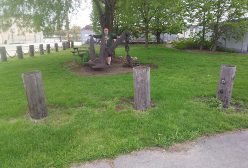 A person stands near a large anchor in a grassy park, surrounded by trees and wooden posts.