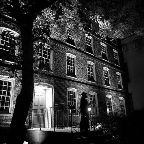 A shadowy figure stands on the steps of a dimly lit brick building at night, surrounded by trees.