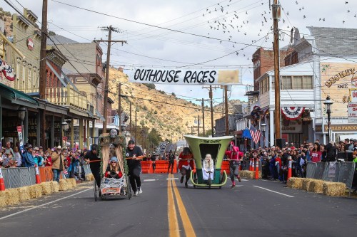 A lively street scene with participants racing in decorated outhouses during a festive event, surrounded by spectators.