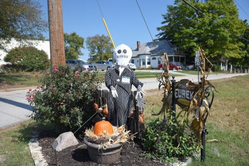 A scarecrow in a striped outfit stands by a pumpkin and autumn decorations near a roadside.